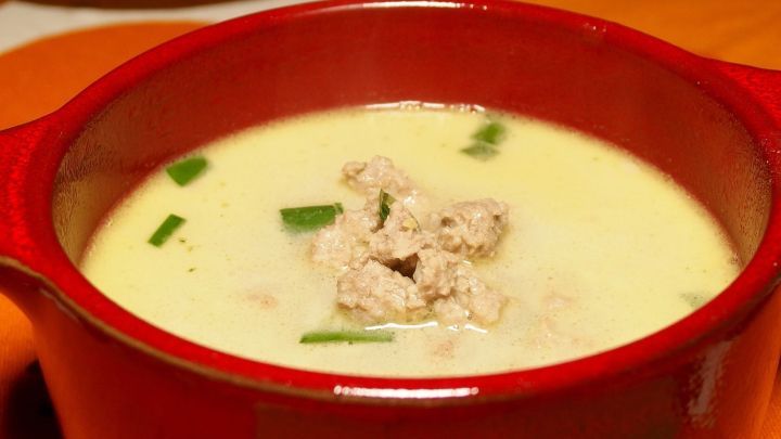 Eine rote Sch&uuml;ssel mit K&auml;sesuppe steht auf einem bunten Tisch. Die Suppe ist cremig, mit Kr&auml;utern und kleinen St&uuml;ckchen dar&uuml;ber.