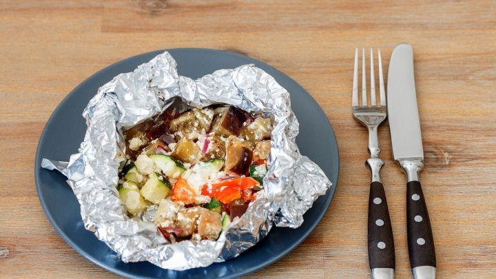 Gem&uuml;se in Alufolie ruht auf einem dunklen Teller; daneben liegen Besteckst&uuml;cke auf dem Holztisch.