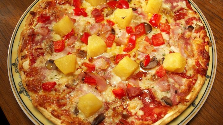 Eine &uuml;ppige Pizza liegt auf einem Holzuntergrund, belegt mit Tomaten, Ananas und Schinken, appetitlich angerichtet.