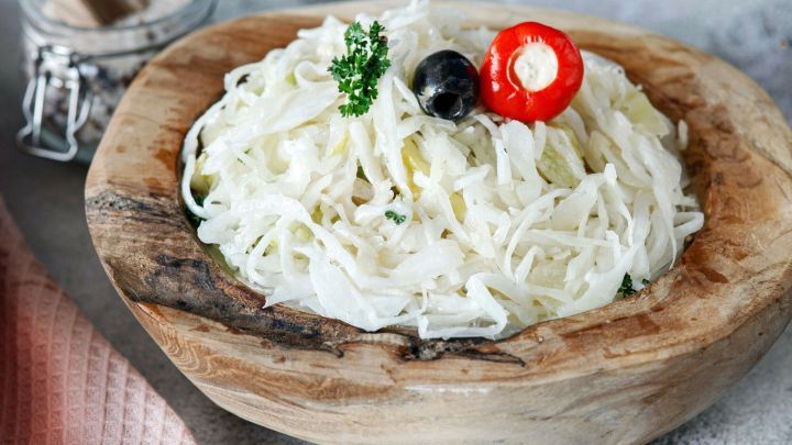Krautsalat lecker serviert in einer Holzsch&uuml;ssel, dekoriert mit Paprika und Oliven auf einem hellen Tisch.