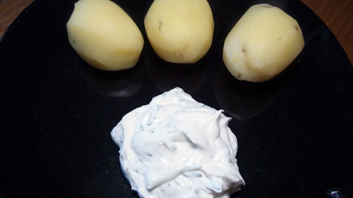 Ein schwarzer Teller mit drei rohen Kartoffeln und einem Klecks Quark als Beilage im Fokus der Darstellung.