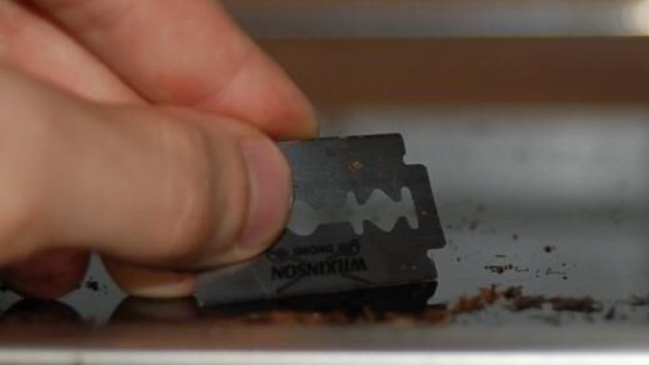 Eine Hand h&auml;lt eine Rasierklinge und s&auml;ubert ein Glaskeramikfeld, indem sie Schmutz von der Oberfl&auml;che kratzt.