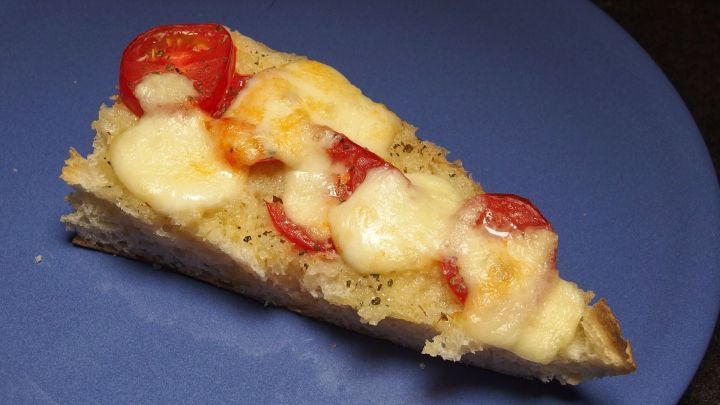 Ein St&uuml;ck Fladenbrot liegt auf einem blauen Teller, &uuml;berbacken mit geschmolzenem K&auml;se und Tomaten, appetitlich angerichtet.