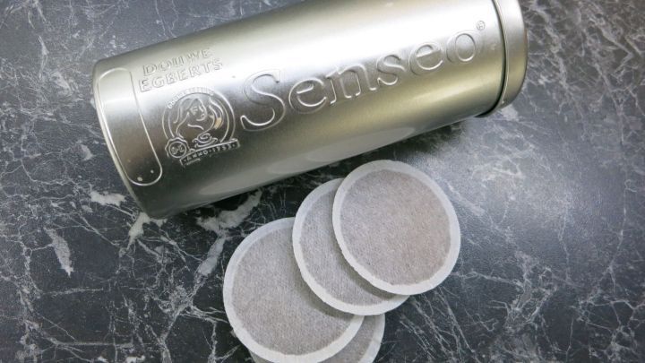Eine silberne Senseo-Dose steht auf einer dunklen Marmoroberfl&auml;che, daneben liegen drei Kaffeepads der dunklen R&ouml;stung.