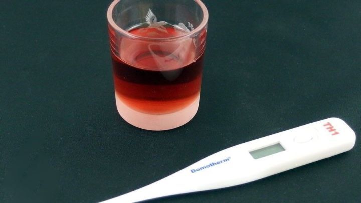 Ein Glas Himbeersaft steht neben einem Fieberthermometer auf einem dunklen Untergrund, ideal gegen Fieber.