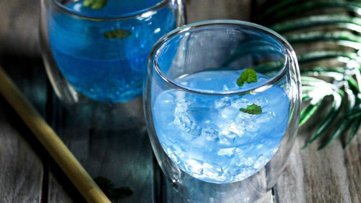 Zwei gl&auml;serne Beh&auml;lter mit blauen Getr&auml;nken und Eisw&uuml;rfeln stehen auf einem Holztisch, umgeben von gr&uuml;nen Bl&auml;ttern.