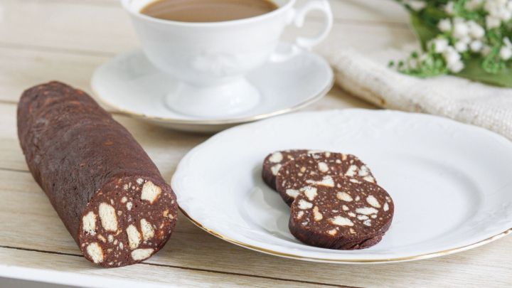 Schoko-Wurst liegt geschnitten auf einem Teller. Daneben steht eine Tasse Kaffee, umgeben von Blumen auf einem Holztisch.