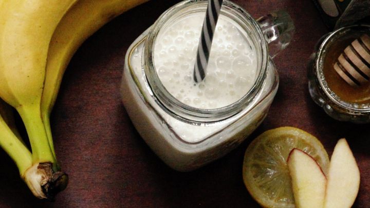 Ein cremiger Banapfel Shake in einem Glas mit Strohhalm steht auf einem Tisch, umgeben von Bananen und Apfelscheiben.
