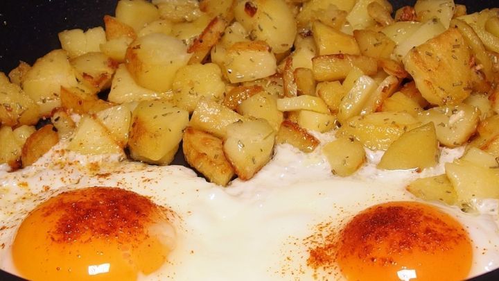In einer Pfanne braten zwei Spiegeleier zusammen mit knusprigen Kartoffelst&uuml;cken, die mit Gew&uuml;rzen verfeinert sind.