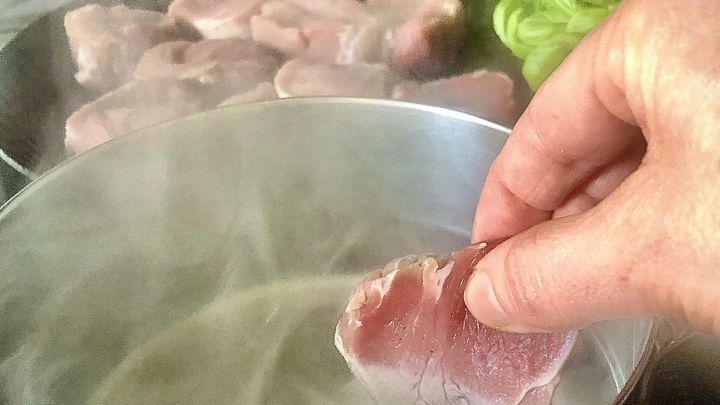 Ein St&uuml;ck Fleisch wird von einer Hand in kochendes Wasser gehoben, w&auml;hrend im Hintergrund weitere Fleischst&uuml;cke dampfen.