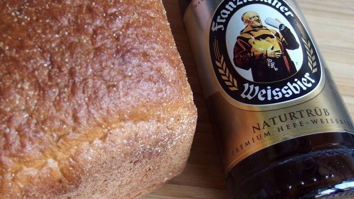 Ein frisches Brot liegt neben einer Flasche Wei&szlig;bier, die als Zutat f&uuml;r das Backen dient, auf einem Holzuntergrund.