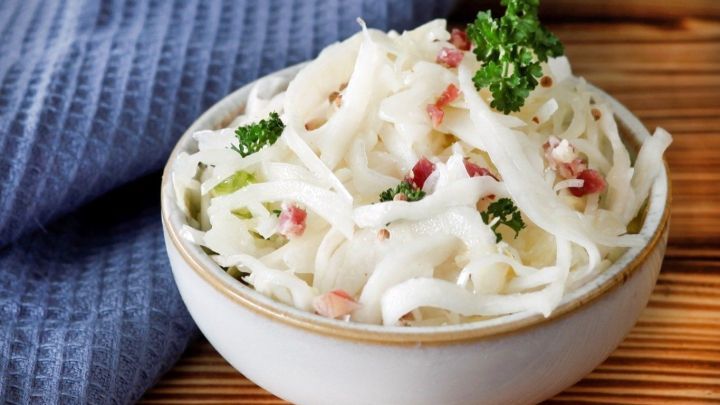 Ein Sch&uuml;ssel mit bayrischem Krautsalat, der frisch zubereitet auf einem Holztisch serviert wird, umgeben von einem blauen Tuch.