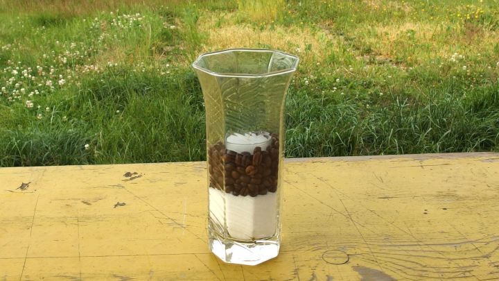 Ein Glas enth&auml;lt geschichtete Kaffeebohnen und Zucker auf einem Holztablett, umgeben von einem gr&uuml;nen Garten.