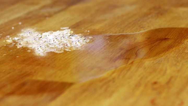 &Ouml;l flie&szlig;t auf einem Holztisch, neben verstreuten Haferflocken, was eine K&uuml;chenreinigungssituation darstellt.