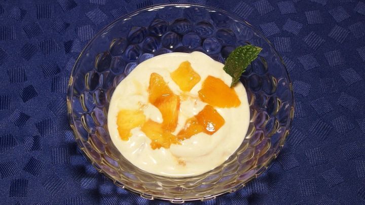 Eine Sch&uuml;ssel Quarkspeise mit Dosenpfirsichen steht auf einem blauen Tisch, dekoriert mit frischen Minzbl&auml;ttern.