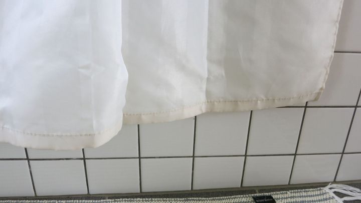 Ein Duschvorhang mit braunem Rand h&auml;ngt vor wei&szlig;en Fliesen und sch&uuml;tzt den Boden beim Duschen.