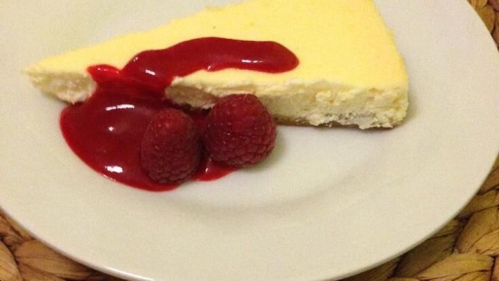 Ein St&uuml;ck K&auml;sekuchen auf einem wei&szlig;en Teller liegt zusammen mit rotem Fruchtso&szlig; und Himbeeren auf einem dekorativen Untersetzer.