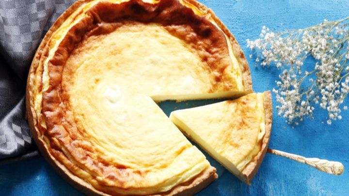 Ein K&auml;sekuchen liegt auf einem blauen Untergrund, mit einem St&uuml;ck herausgeschnitten und einer Gabel daneben.