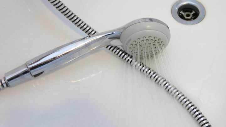 Eine Duschbrause spritzt Wasser auf eine Badewanne, w&auml;hrend der Abfluss sichtbar im Hintergrund zu sehen ist.