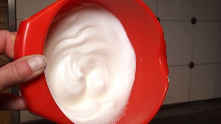 Eine Hand h&auml;lt eine rote Sch&uuml;ssel mit steif geschlagenem Eiwei&szlig;, umgeben von einer modernen K&uuml;chenumgebung.