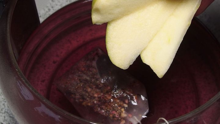 Ein Teebeutel wird in eine tiefrote Fl&uuml;ssigkeit eingelegt, w&auml;hrend ein Apfelst&uuml;ck &uuml;ber dem Getr&auml;nk schwebt.