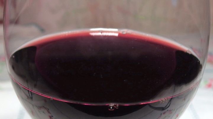 Ein Weinglas steht auf einem Tisch und enth&auml;lt tiefroten Rotwein. Im Hintergrund sind verschwommene Tapetenmuster zu sehen.