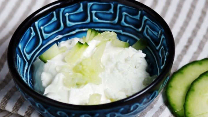 Ein blauer Schalen voll leckerem selbstgemachten Tsatsiki steht auf einem gestreiften K&uuml;chentuch, umgeben von Gurkenscheiben.