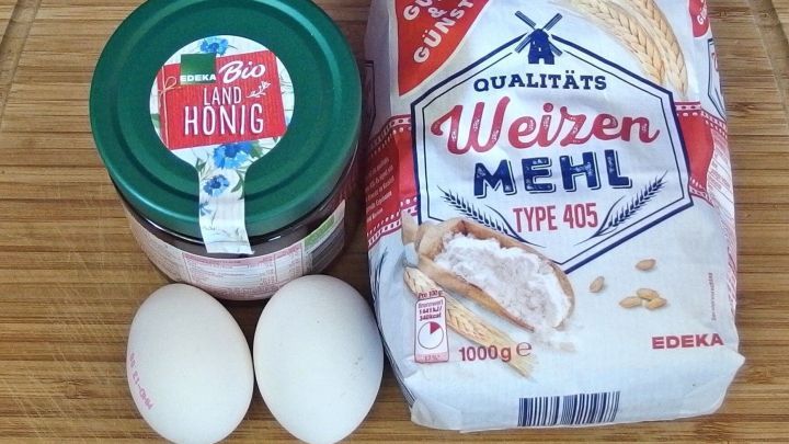 Zwei Eier, ein Honigglas und eine Packung Weizenmehl liegen auf einem Holzbrett, bereit zur Verwendung in einer Gesichtsmaske.