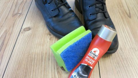 Ein Küchenschwamm liegt neben einer Flasche Schuhcreme und schwarzen Schuhen auf einem Holzfußboden, bereit zum Eincremen.