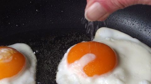 Zwei Spiegeleier braten in einer Pfanne, während jemand Salz über das Eigelb streut, um sie richtig zu würzen.