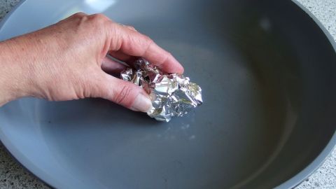 Eine Hand hält ein Stück Alufolie, das in einer grauen Pfanne mit Wasser verwendet wird, um die Pfanne zu reinigen.