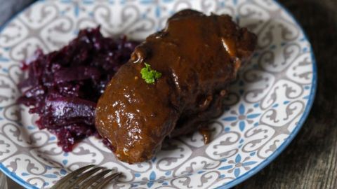 Die Zubereitung von Rouladen ist ohne lästige Spieße und Zahnstocher mithilfe von diesem Tipp möglich. Auf einem dekorativen Teller liegt eine saftige Roulade, umgeben von Rotkohl. Die Roulade glänzt und ist appetitlich angerichtet.