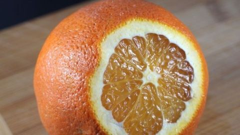 Apfelsinen vor dem Schälen kurz in heißes Wasser legen, dann lassen sie sich leichter schälen. Eine orangefarbene Orange ist auf einem Holzbrett in der Mitte durchgeschnitten, wobei die fruchtigen Segmente sichtbar sind.