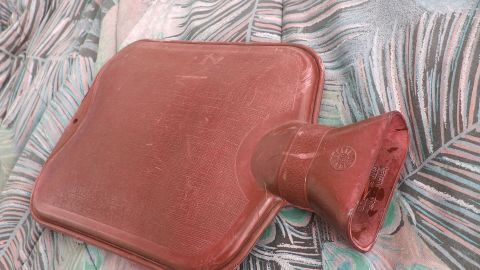 Eine rote Wärmflasche liegt auf einem bunten, gemusterten Untergrund und ist bereit, länger warmes Salz zu halten.