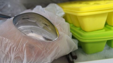 Eine Hand mit einem Löffel, eingehüllt in eine Plastikhandschuh, greift nach einem Gelbeisbehälter im Gefrierfach.