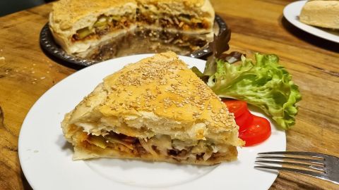 Eine Stück Fladenbrotpizza liegt auf einem Teller und wird von frischem Salat sowie Tomaten umgeben, während im Hintergrund eine große Pizza sichtbar ist.