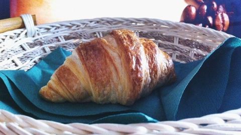 Ein frisch gebackenes Croissant liegt auf einem teal-farbenen Tuch in einem Korb, umgeben von ansprechendem, weichem Licht.