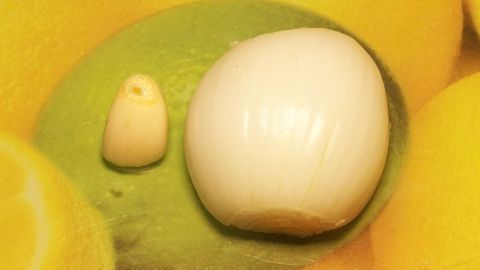 Mit Minzpaste Zwiebel- und Knoblauchgeruch an den Händen entfernen. Eine weiße Zwiebel und eine Knoblauchzehe liegen auf einer grünen Unterlage, umgeben von gelbem Hintergrund.