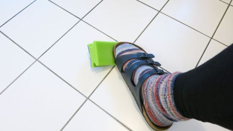 Ein Fuß mit einem bunten Socken drückt auf einen grünen Wischmop auf einem glatten Boden, während der Raum hell erleuchtet ist.