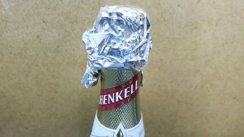 Verwende Alufolie, um geöffnete Sektflaschen frisch zu halten. Einfach den Flaschenhals fest umwickeln. So bleibt die Kohlensäure länger erhalten und der Sekt genießbar. Ideal für spontane Partys und gesellige Abende. Eine Sektflasche mit Aluminiumfolie als Verschluss bedeckt, steht vor einem neutralen Hintergrund. Text: „HENKELL“.