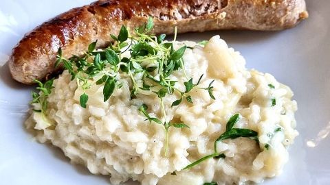 Cremiges Risotto mit frischem Thymian trifft auf knusprige Bratwurst – eine köstliche Variation, die in der Artikelreihe "Reisvariationen" ins Rampenlicht gerückt wird. Perfekt für ein herzhaftes und aromatisches Gericht. Würzige Bratwurst liegt neben cremigem Risotto mit Kräutern garniert auf einem weißen Teller, ready to enjoy.
