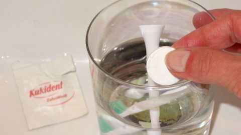 Die Aufsätze von elektrischen Zahnbürsten lassen sich hervorragend mit Gebissreiniger (Kukident) reinigen. Eine Hand gibt eine Reinigungstablette in ein Glas Wasser, das einen Zahnbürstenaufsatz mit Gebissreiniger enthält.