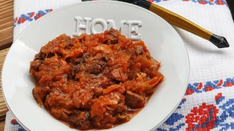 Ein weißer Teller mit polnischem Bigos steht auf einem dekorierten Tuch. Ein Löffel liegt daneben.