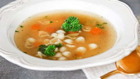Eine Tütensuppe in einer weißen Schüssel, gefüllt mit Brühe, Nudeln und Gemüse, steht auf einem stoffartigen Untergrund.