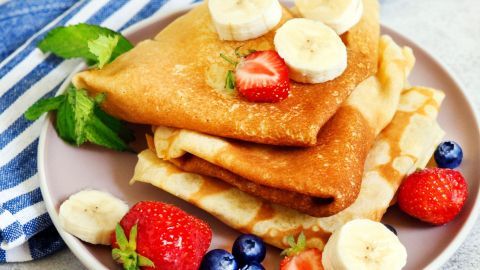 Goldbraune vegane Pfannkuchen liegen auf einem Teller, garniert mit Bananen, Erdbeeren und blauen Beeren, umgeben von frischen Minzblättern.