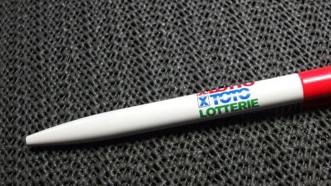 Ein roter Kugelschreiber mit einer weißen Hülse liegt auf einem strukturierten schwarzen Stoff. Aufschrift: "Lotto".