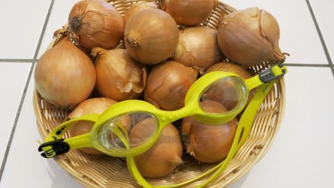 In einem Korb liegen mehrere Zwiebeln zusammen mit einer gelben Schutzbrille, die humorvoll gegen das Weinen beim Schneiden schützt.