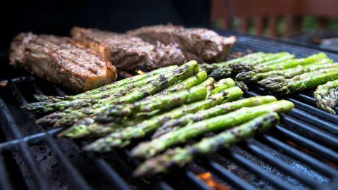 Spargel liegt auf einem Grill und wird geröstet, während nebenan Fleisch brät; ein schönes Grillambiente im Freien.