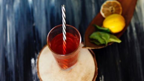 Ein Glas mit rotem Getränk und gestreiftem Strohhalm steht auf einem Holzbrett. Daneben liegen Zitrone und Limettenblätter.