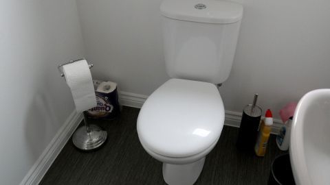 Eine moderne Toilette steht auf einem schlichten, grauen Boden, umgeben von einfachen Reinigungsmitteln und Papier.
